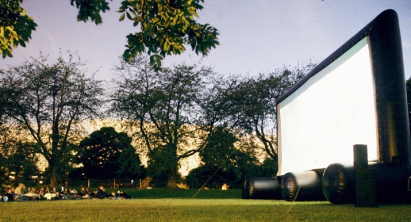 People watching an outdoor pop-up cinema screen in a park at dusk