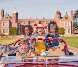 Four children dressed as characters from Wind in the Willows in an old fashioned car infront of an English stately home