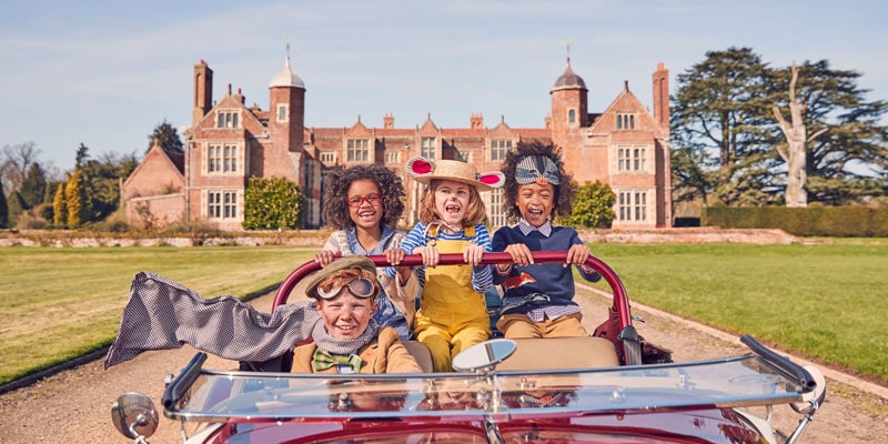 Four children dressed as characters from Wind in the Willows in an old fashioned car infront of an English stately home
