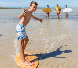 A boy surfing the shallows on the beach at Agon-Coutainville, Normandy with his family in the water behind