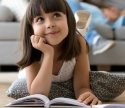 Close up little adorable thoughtful smiling daughter dreaming lying at cushion on warm floor with book in living room at modern home, resting married couple parents on background, focus on small kid (Close up little adorable thoughtful smiling daughte