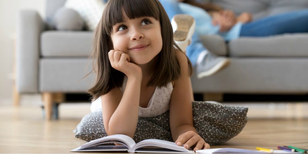 Close up little adorable thoughtful smiling daughter dreaming lying at cushion on warm floor with book in living room at modern home, resting married couple parents on background, focus on small kid (Close up little adorable thoughtful smiling daughte